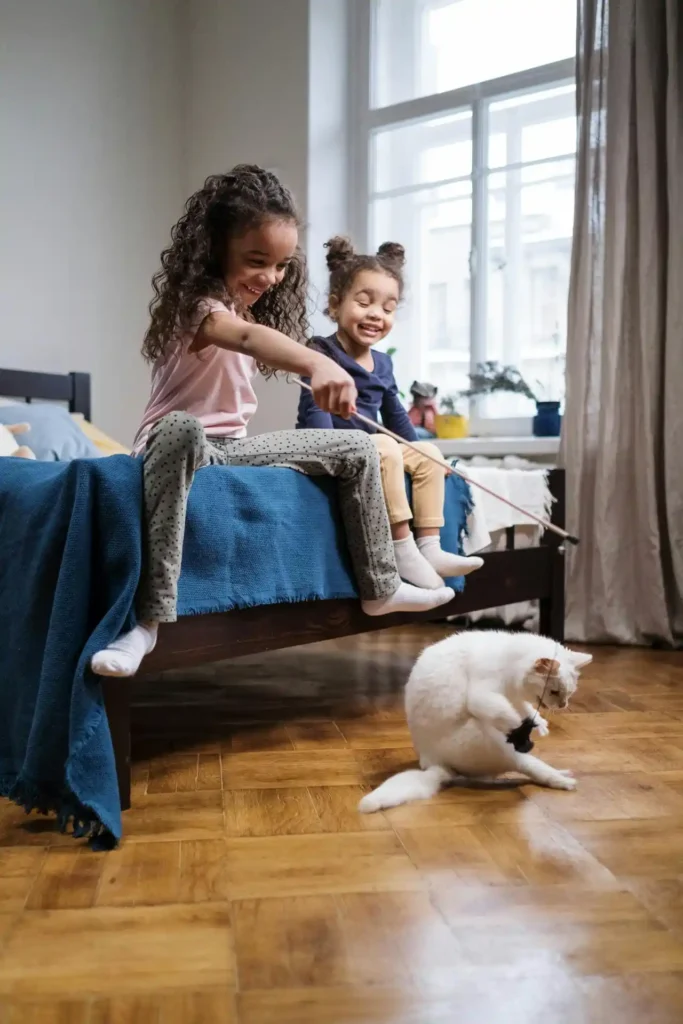 Brincadeiras para Gatos: 4 Temas Importantes seu Felino 7 Crianças brincando com um gato branco usando um brinquedo de varinha em um quarto